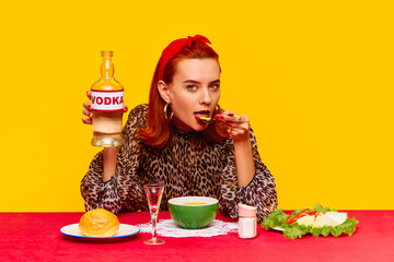 Beautiful stylish redhead woman sitting at table against yellow background and having dinner with cheese soup and drinking vodka. Food pop art photography. Complementary colors. Copy space for ad