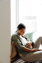 Pregnant woman caressing her belly while sitting on windowsill at home