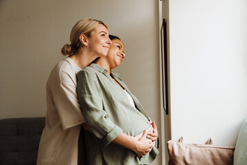 Pregnant lesbian couple hugging and looking through window while standing together at home