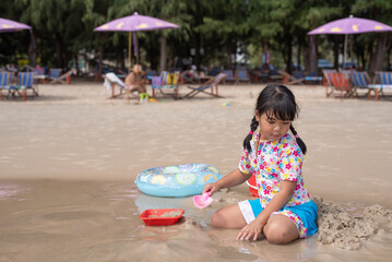 Front view portrait with copy space of 6-year-old preschool Asian girl sitting at beach on a sunny day, playing alone, building sand castle with a plastic toy, wearing a swimming suit. Kid concept