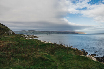 Path Glenarm Coastal Path, south of the village of Glenarm, in Northern Ireland, landscape with mountains, sea, grass and cloudy blue sky