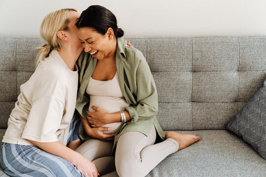 Woman kissing her pregnant wife while sitting on sofa