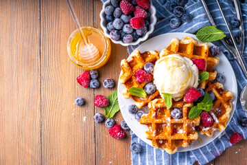 Tasty Homemade Belgian waffles with raspberries, blueberries, vanilla ice cream and syrup or honey, sweet summer healthy breakfast, light wooden background copy space 