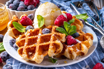 Tasty Homemade Belgian waffles with raspberries, blueberries, vanilla ice cream and syrup or honey, sweet summer healthy breakfast, light wooden background copy space 