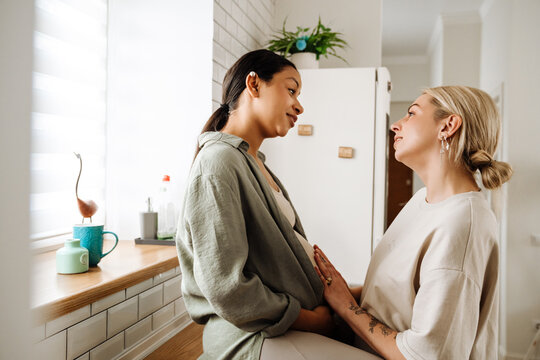 Woman smiling and touching belly of her pregnant wife while spending time together in kitchen