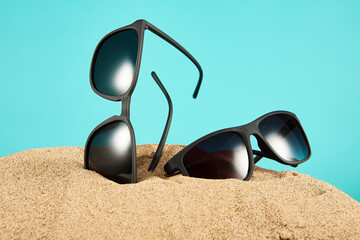 Pair of black sunglasses vertically sticking out of sand on blue.