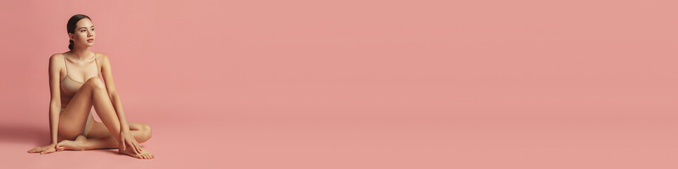 Fototapeta premium One young beautiful tanned girl with perfect figure in underwear lying on floor over pink studio background. Concept of female beauty, tenderness, body and skin care