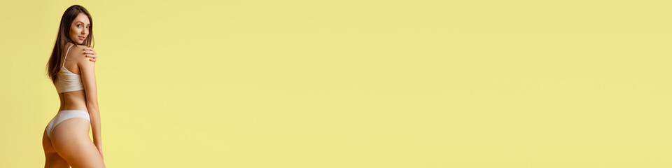 Fototapeta premium Pretty young girl posing in cotton underwear over yellow studio background. Weight loss. Concept of beauty, body and skin care, fitness, health, spa, cosmetics, ad