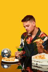Food pop art photography. Young man in stylish shirt sitting at table, drinking cocktail and eating pizza against yellow studio background. Party. Complementary colors. Copy space for ad, text