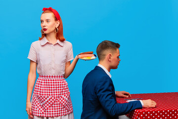 Emotionaless. Young redhead woman serving breakfast with fried eggs and sausages to man against blue studio background. Food pop art photography. Complementary colors. Copy space for ad, text
