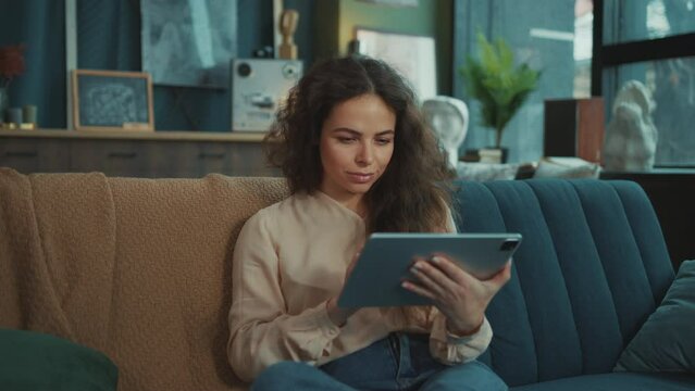 Portrait of attractive stylish girl holding digital tablet in hands, scrolling through screen while working at home. Young beautiful Caucasian woman looking directly at camera. Freelance concept