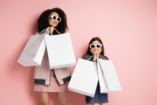 Fashionable mother and daughter in sunglasses holding shopping bags on pink background.
