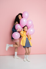 Stylish woman and daughter looking at camera near balloons on pink background.