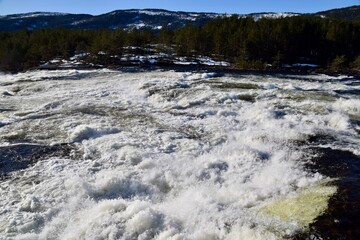 Fototapeta premium White Water rushing over rocks with Trees and hills and blue skies behind. Byglandsfjord, Norway, Feb 2023. 