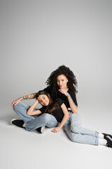 Tattooed woman and daughter in jeans and t-shirts smiling at camera on grey background.