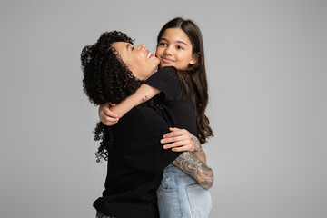 Cheerful girl in jeans and t-shirt hugging tattooed mom isolated on grey.