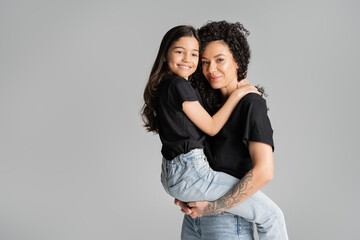 Curly woman lifting preteen daughter and looking at camera isolated on grey.