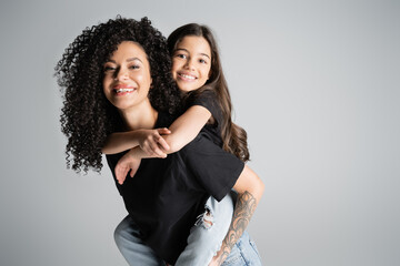 Cheerful girl piggybacking on curly tattooed mom isolated on grey.