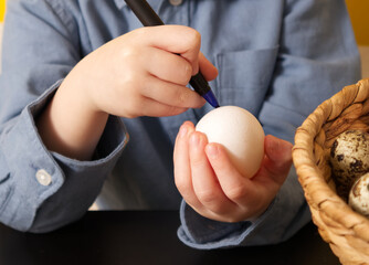 The hands of a little boy hold a chicken egg and a felt-tip pen and paint an easter egg close up