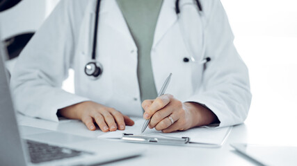 Unknown doctor woman sitting and writing notes at the desk in clinic or hospital office, close up.  Medicine concept.