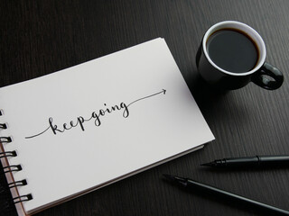 KEEP GOING letternig with arrow showing the way forward in notebook with cup of coffee and pens on black wooden desk