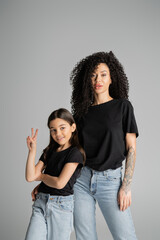 Preteen kid showing peace sign near stylish mom isolated on grey.