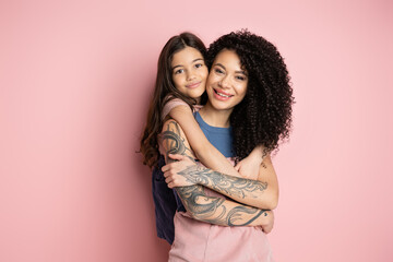 Smiling preteen girl hugging tattooed mother on pink background.