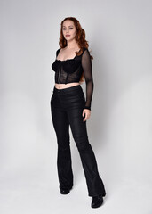 Fototapeta premium Full length portrait of beautiful woman with long red hair wearing black corset top and leather pants. Standing pose, walking forwards with gestural hands reaching out. Isolated on white studio backgr