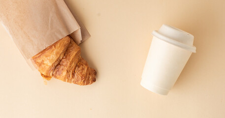 Top view of freshly baked croissant with golden crust next to a disposable paper cup of coffee. French traditional pastry on the beige background. Breakfast in the morning concept