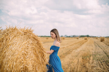 Naklejka na ściany i meble Portrait of a beautiful woman in a summer dress on a field among haystacks in summer.