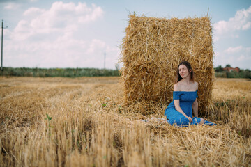 Naklejka na ściany i meble Portrait of a beautiful woman in a summer dress on a field among haystacks in summer.