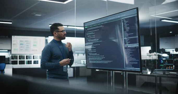 Handsome Multiethnic Lead Software Engineer Standing Alone in a Meeting Room, Using a Big TV Display to Review a Computer Program Code Created by Research and Development Department