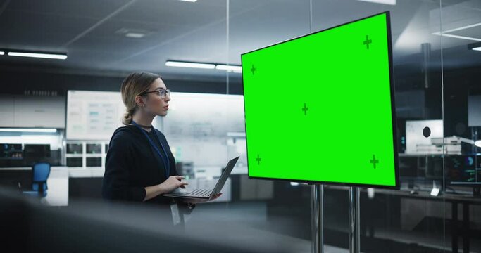Portrait of a Multiethnic QA Engineer Working on Finding and Fixing Bugs in a Product or Software Before the Launch. Female Using Laptop in a Room with Big Green Screen Mock Up Display