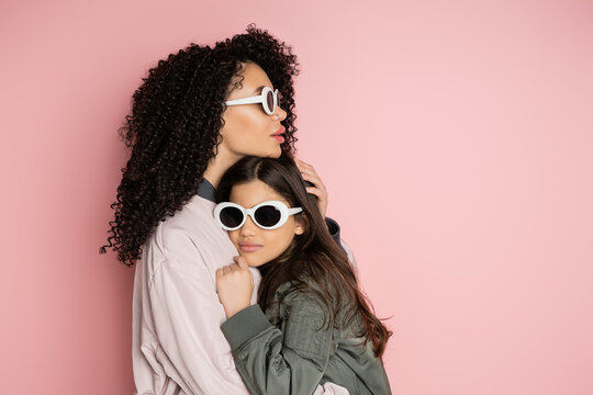 Stylish woman hugging daughter in sunglasses on pink background.