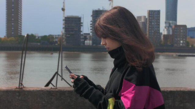 Side view of a young woman walking near a river in the city while using her mobile phone