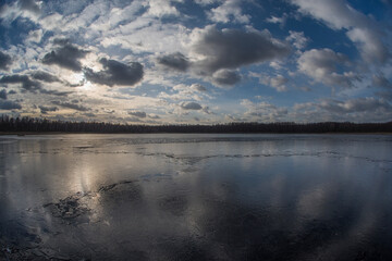 Fototapeta na wymiar Evening landscape of a spring lake covered with thin cracking transparent ice and a cloudy sky reflected in it. Fish eye.