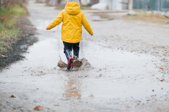 A child in a yellow raincoat and rubber boots runs through the puddles. View from the back, unrecognizable figure. Little boy plays in the water 2-3 years old