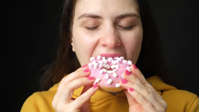 A woman licks looking at a pink marshmallow donut and bites it, a black background.
