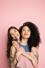 Smiling girl sticking out tongue while hugging curly mother on pink background. - obrazy, fototapety, plakaty