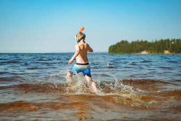 Fototapeta premium cute caucasian boy wearing snorkel mask running into water with splashes. Vacation on sea side.