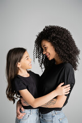 Side view of positive woman and daughter in black t-shirts hugging isolated on grey.