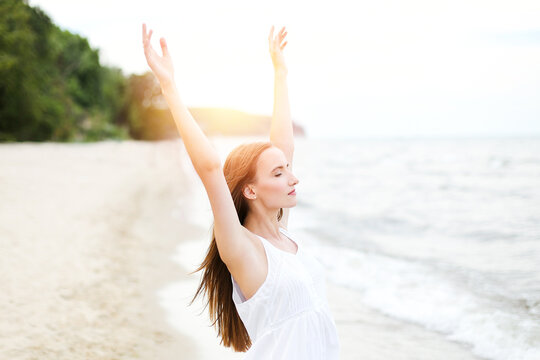 Happy smiling woman in free happiness bliss on ocean beach standing with raising hands. Portrait of a multicultural female model in white summer dress enjoying nature