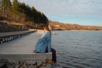 Young woman sits on the shore of the lake, enjoying in her free time.