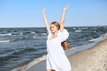 Happy smiling beautiful woman on the ocean beach standing in a white summer dress, raising hands.
