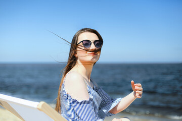 Happy young brunette woman relaxing on a wooden deck chair at the ocean beach while smiling and wearing fashion sunglasses. The enjoying vacation concept.