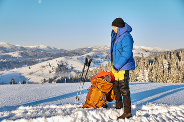 Tourist travelling in winter dressed in winter sportswear with backpack walking with trekking poles in the snowy pine mountains. Traveller with his backpack looking on the hill. 