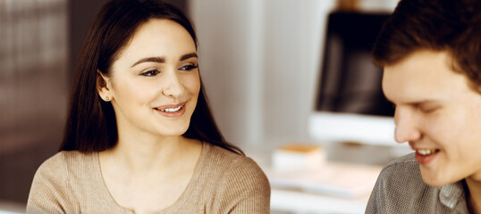 Friendly young businessman and programmer in a green shirt is working on computer, while sitting together at the desk with a female colleague in a modern office. Focus on woman