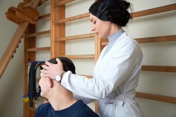 Young attractive female rehabilitation doctor in a white medical suit is working on the recovery of a young sportsman wrestler after a neck injury. Office of physical rehabilitation.