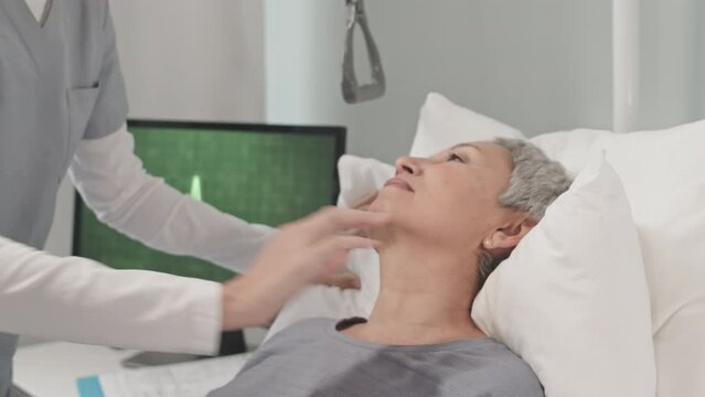 Unrecognizable caring nurse helping senior Asian woman lying on bed in hospital room, laying soft pillow under patient back to feel more comfortable