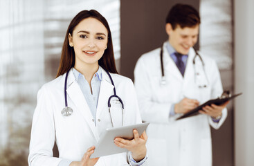 Fototapeta premium Smiling intelligent woman-doctor is holding a tablet computer in her hands, while she is standing in a clinic with a colleague at the background. Portrait of physician at work